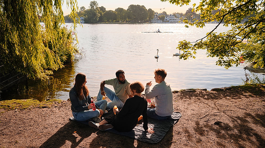 Gruppe junger Menschen sitzt entspannt am Alsterufer in Hamburg bei Sonnenuntergang mit Blick aufs Wasser