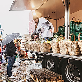 Händler auf dem Hamburger Fischmarkt verkauft Körbe mit Waren vom Wagen, Besucher:innen mit Regenschirmen