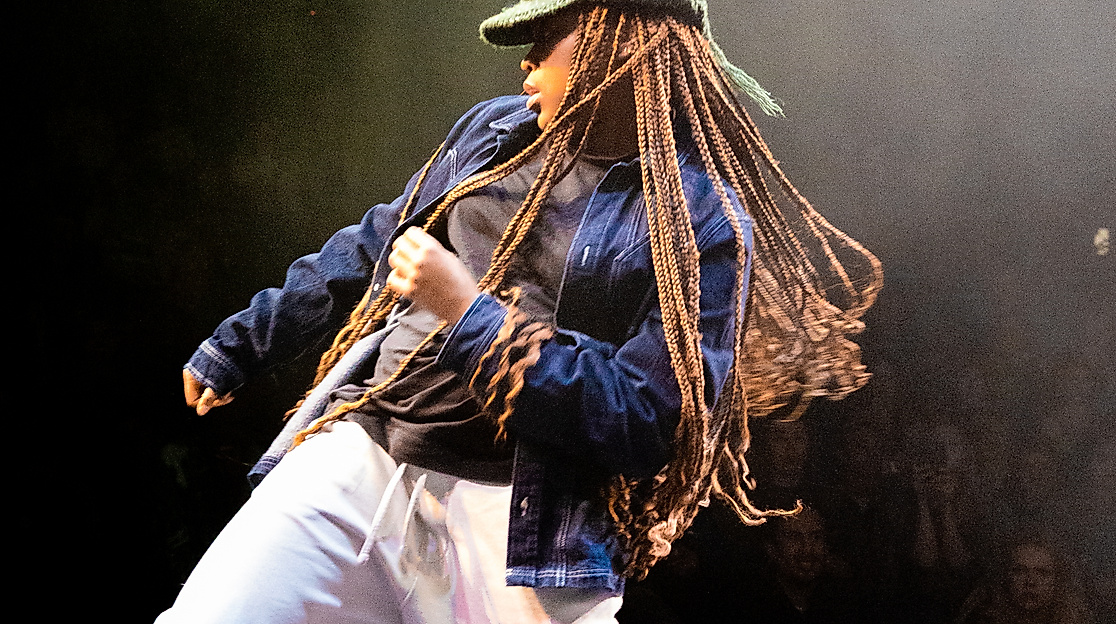 A person wearing a knitted hat, long braids, and loose clothing stands on a stage and moves their hips in one direction with bent knees. They pull themselves up in the same direction with the opposite arm.