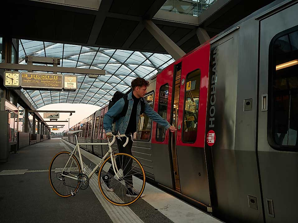 Ein junger Mann mit Rucksack und Fahrrad steigt an einem überdachten S-Bahnhof in eine rote S-Bahn mit der Aufschrift „HOCHBAHN“ ein. Die Szene zeigt moderne, umweltfreundliche Mobilität in Hamburg – flexibel kombinierbar mit Fahrrad und öffentlichem Nahverkehr. Ideal für Entdeckungen mit der Hamburg CARD, die freie Fahrt im HVV bietet.
