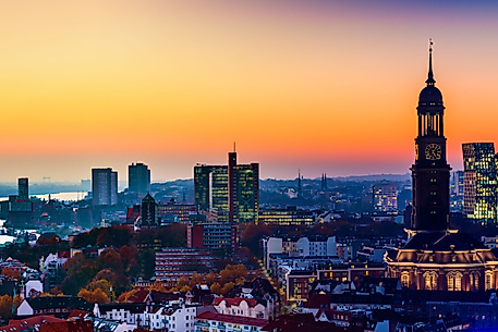 Panorama von Hamburg bei Sonnenuntergang mit beleuchtetem Michel-Turm und Hafen im Hintergrund.