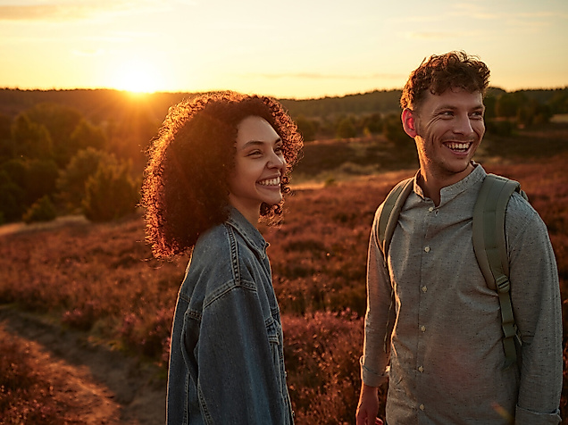 A young man and woman stand laughing in the sunset in the Lüneburg Heath, which is blooming purple.