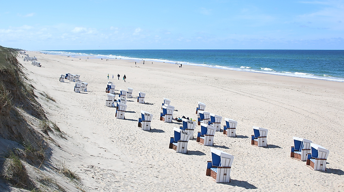 Breiter Sandstrand mit Strandkörben und Spaziergänger:innen an der Nordsee bei Sonnenschein.