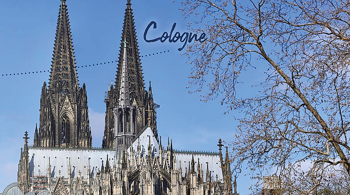 View of Cologne Cathedral’s twin spires against a clear blue sky with tree branches and the word “Cologne” above.