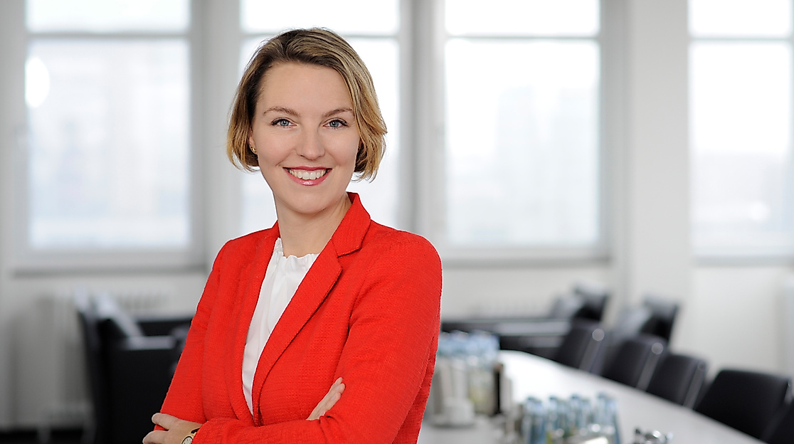 Porträt einer lächelnden Frau im roten Blazer vor einem hellen Konferenzraum, Ansprechpartnerin der Hamburg Tourismus GmbH