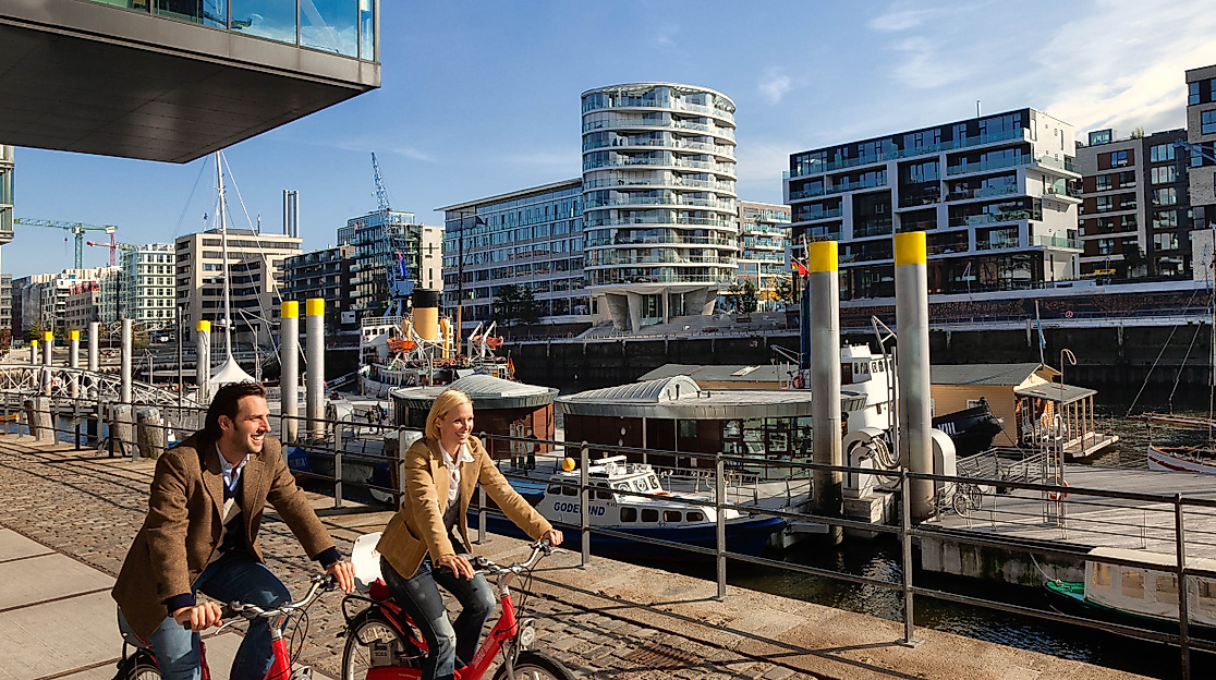 Zwei Personen fahren mit Stadträdern am Sandtorkai in der HafenCity Hamburg bei Sonnenschein entlang des Hafens