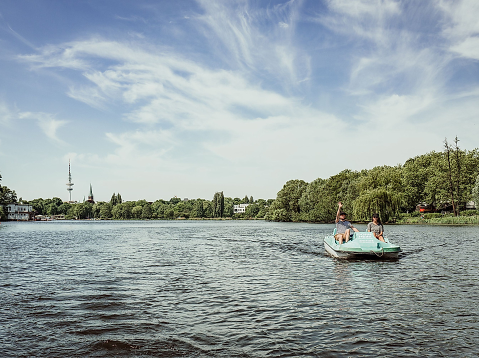 Menschen auf einem Tretboot bei Sonnenschein auf der Alster, umgeben von grüner Uferlandschaft