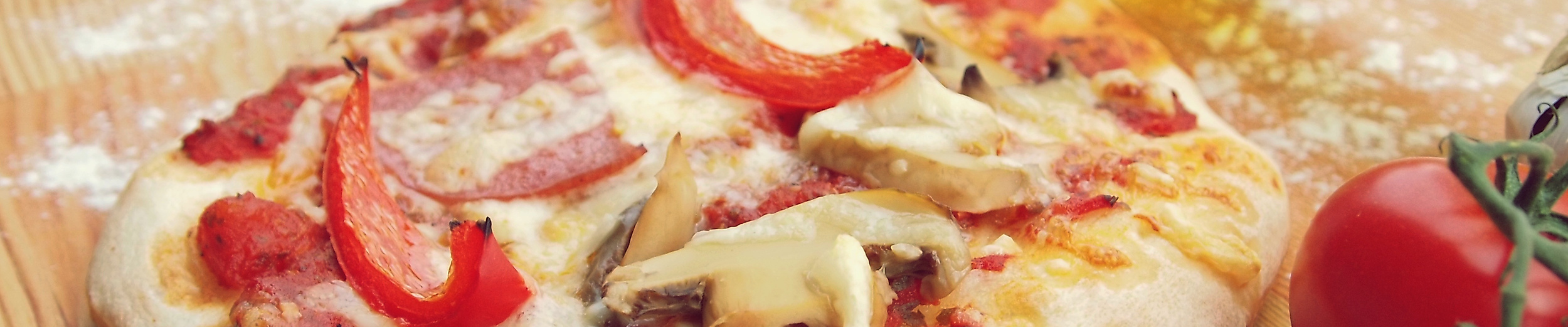 Frisch gebackene Pizza mit Paprika, Pilzen und Schinken auf Holzbrett, daneben Tomaten und Pizzaschneider.