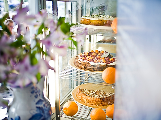 Kuchenvitrine mit frischen Torten und Orangen im Café Johanna, daneben Frühlingsblumen in einer Vase
