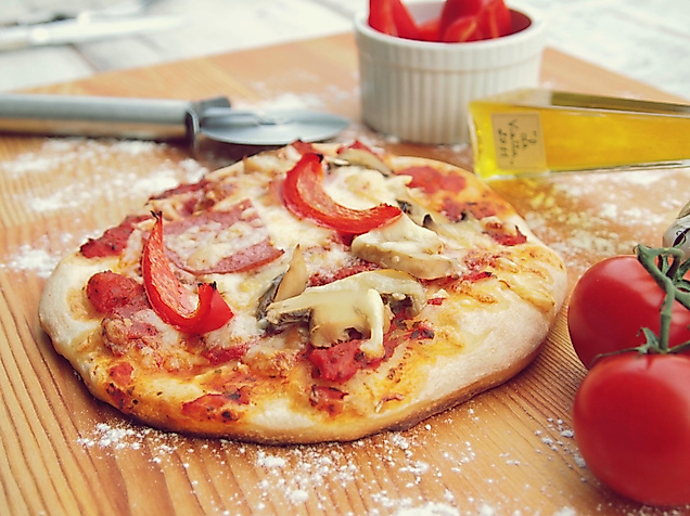 Frisch gebackene Pizza mit Paprika, Pilzen und Schinken auf Holzbrett, daneben Tomaten und Pizzaschneider.