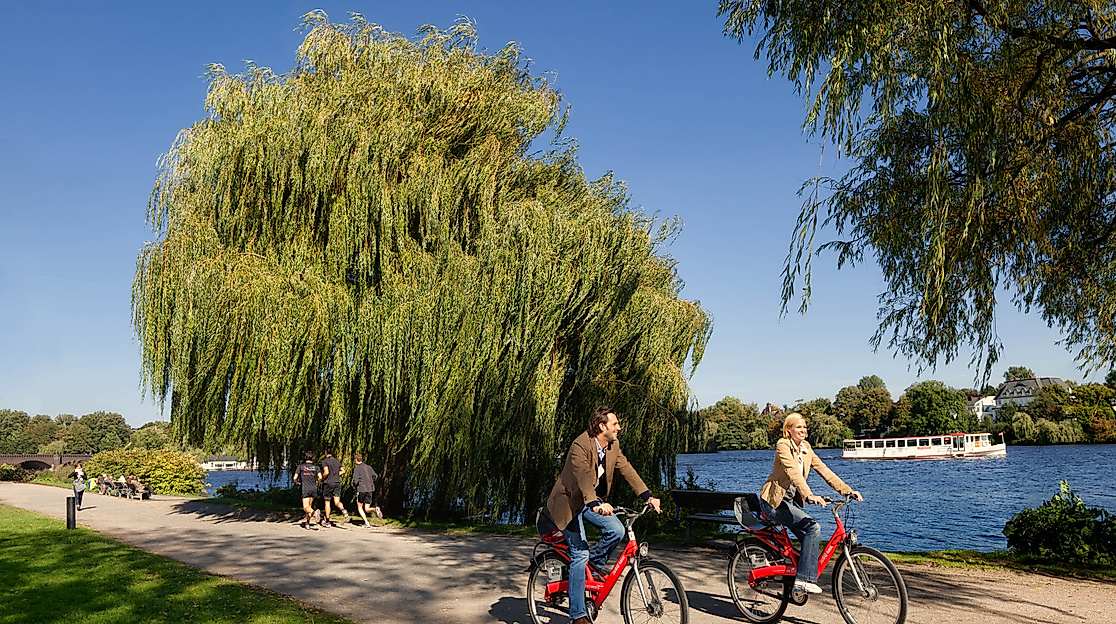 Zwei Personen radeln mit Stadträdern am Ufer der Alster entlang unter blauem Himmel.