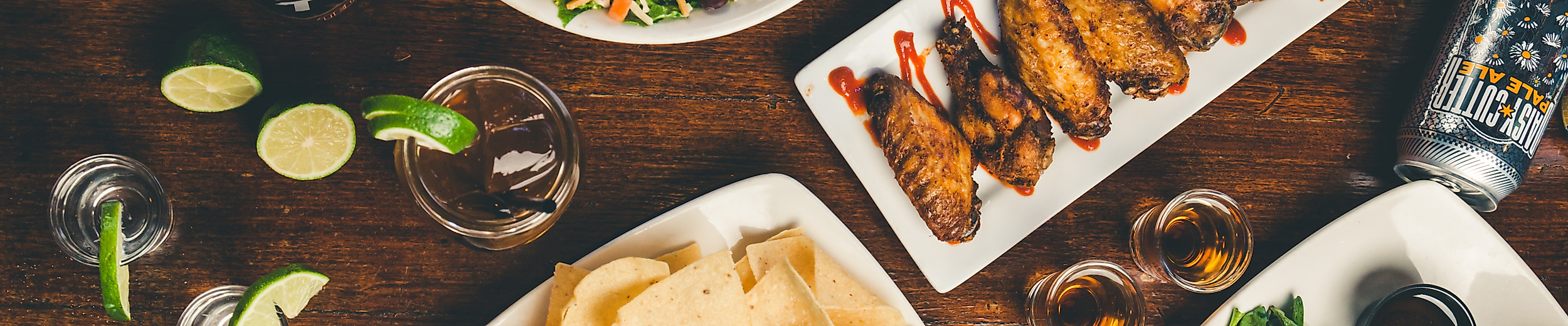 Große Auswahl an Speisen wie Salat, Chicken Wings, Hummus mit Brot und Burger auf Holztisch angerichtet.