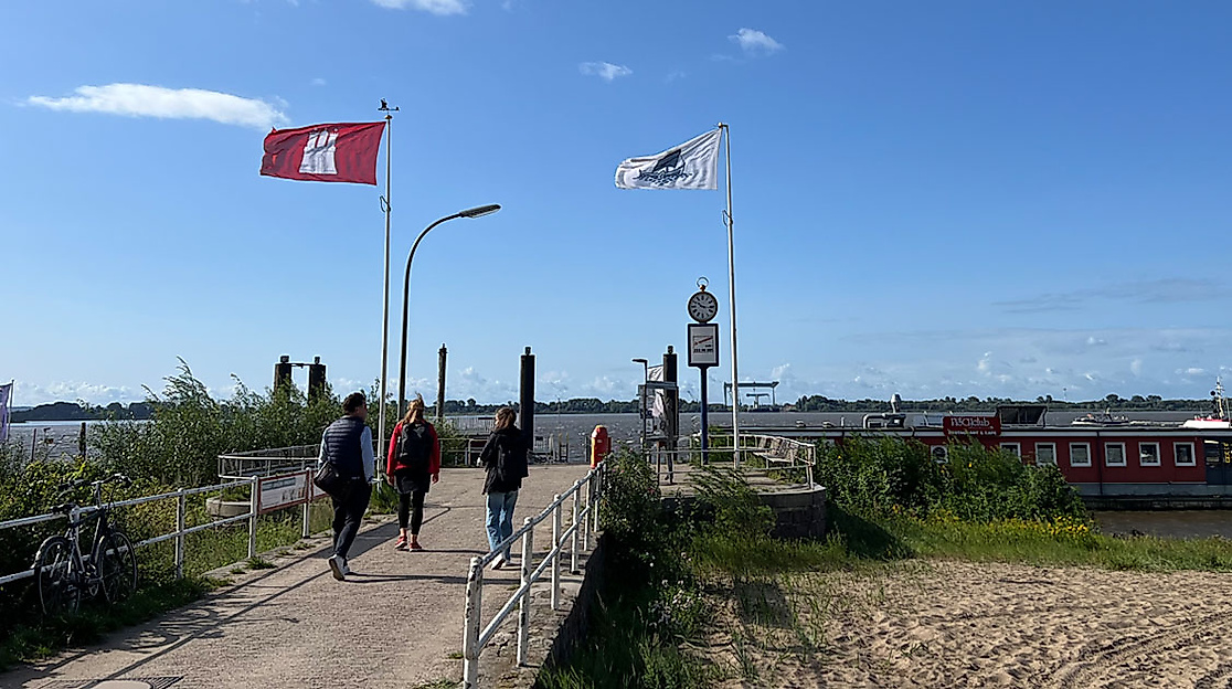 Fähranleger in Blankenese mit wehenden Flaggen und Spaziergänger:innen am Elbufer bei Sonnenschein