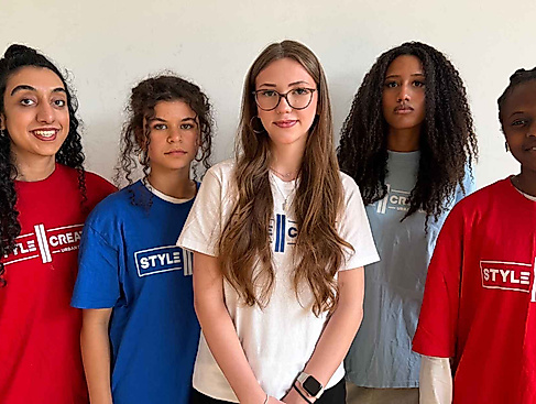 Five people with different hair lengths and textures are wearing colorful T-shirts with the words “STYLE CREATE” printed on them and are standing in front of a light-colored wall.
