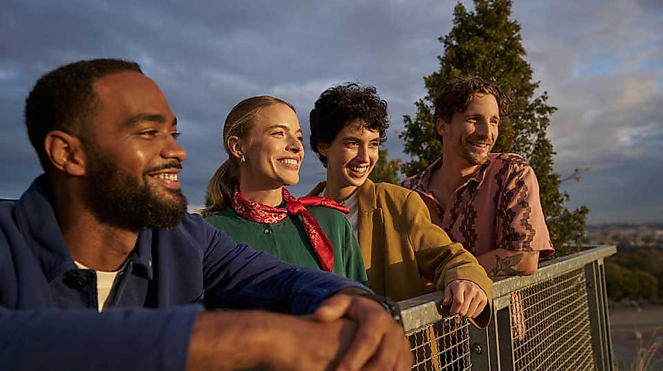 Vier junge Erwachsene stehen entspannt an einem Geländer und blicken lächelnd in die Ferne. Die Szene spielt im warmen Licht eines Sonnenuntergangs, im Hintergrund sind Bäume und ein weiter Blick über die Stadt Hamburg zu erkennen. Die Gruppe wirkt gelöst und fröhlich – ein Moment gemeinsamer Entdeckung und Lebensfreude
