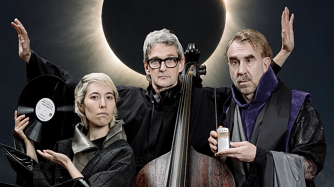 Three people wearing dark, futuristic clothing are posing in front of a solar eclipse.