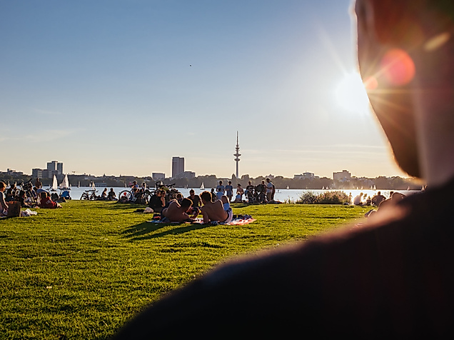 Menschen entspannen im Alsterpark bei Sonnenschein mit Blick auf die Außenalster und die Skyline Hamburgs