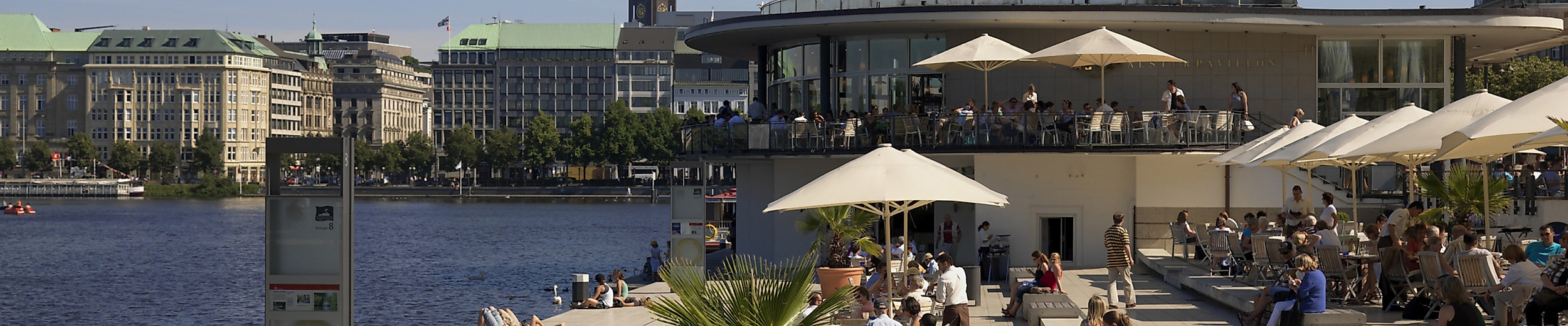 Menschen entspannen am Alsterpavillon in Hamburg bei Sonnenschein, Blick auf die Binnenalster und historische Altstadtfassaden.